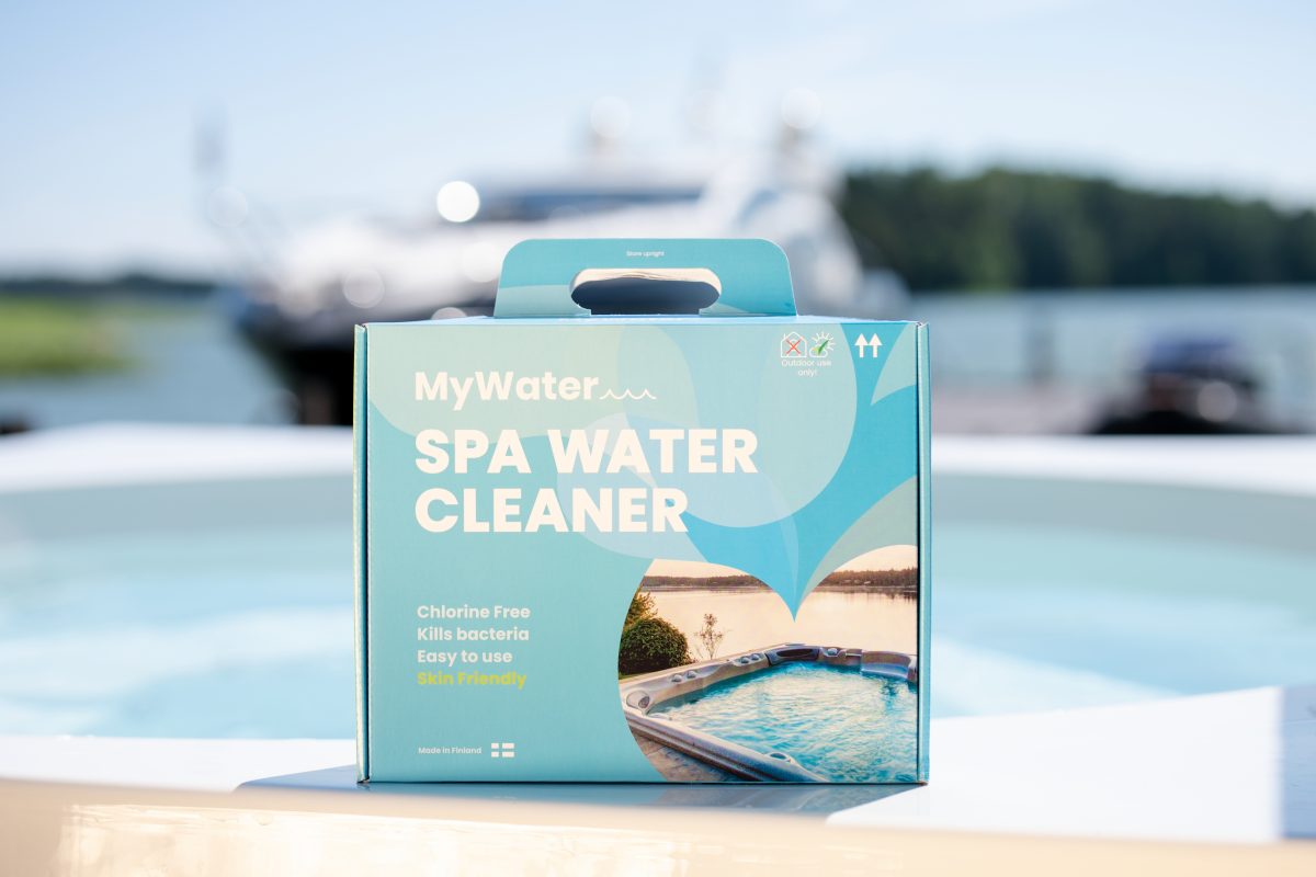 En MYWATER SPA WATER CLEANER-låda på kanten av bubbelbadet, med vatten och båtar i bakgrunden.